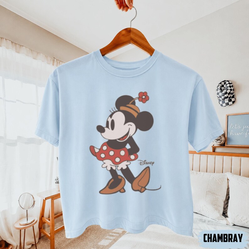 May include: Light blue t-shirt featuring a vintage-style illustration of Minnie Mouse. Minnie is depicted in a red polka-dot dress, brown shoes, and a hat with a flower. The Disney logo is visible. The shirt is hanging on a wooden hanger.