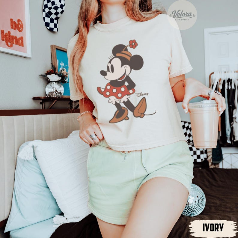 May include: Ivory-colored t-shirt featuring a Minnie Mouse graphic. The cartoon character is depicted in a red polka-dot skirt, black top, and brown shoes. The shirt is paired with light green shorts. The word "Disney" is printed on the shirt.