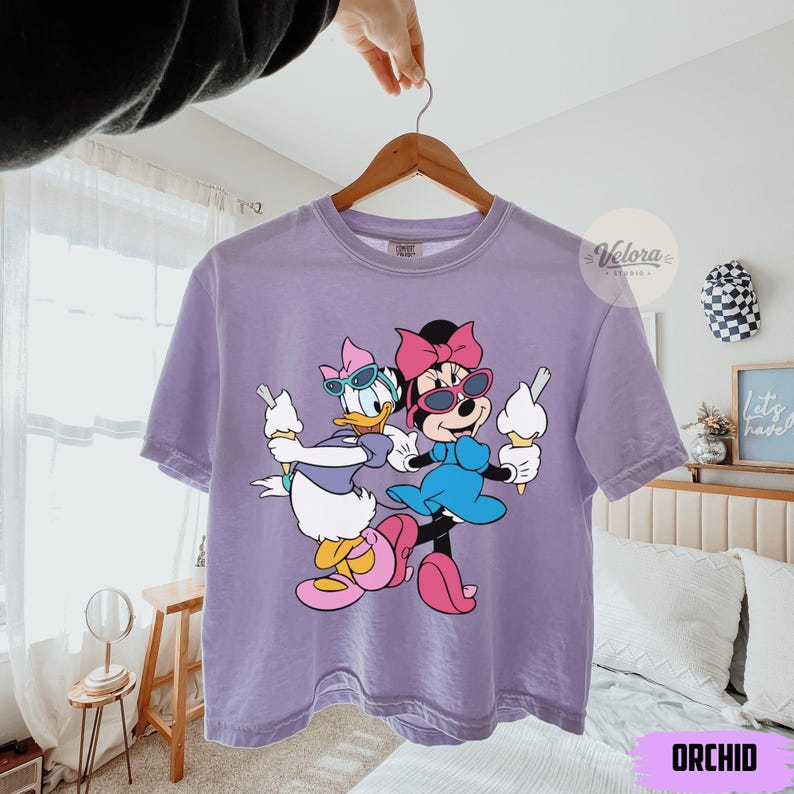 May include: Orchid-colored t-shirt featuring a graphic of Minnie Mouse and Daisy Duck holding ice cream cones. The cartoon characters wear sunglasses and colorful outfits. The shirt is hanging on a wooden hanger.