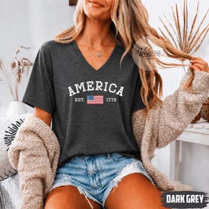 May include: Dark gray V-neck t-shirt with the word "AMERICA" in white block letters above a small American flag and the year "1776". The shirt is made of a soft material and is paired with denim shorts.
