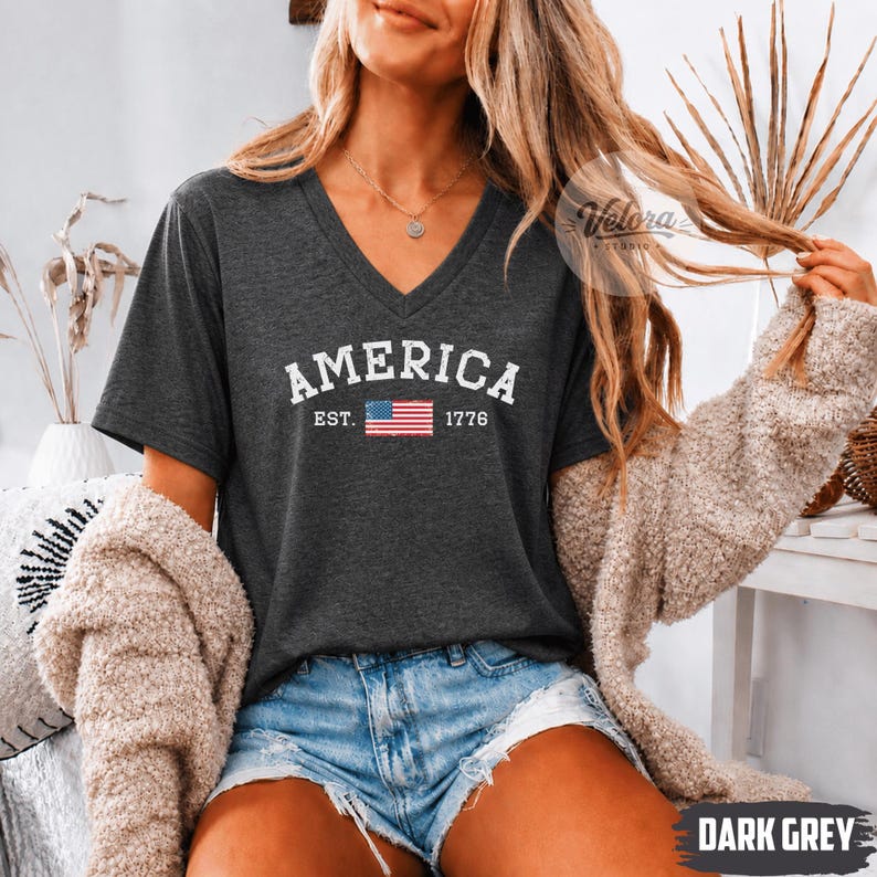 May include: Dark gray V-neck t-shirt with the word "AMERICA" in white block letters above a small American flag and the year "1776". The shirt is made of a soft material and is paired with denim shorts.