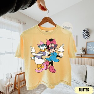 May include: Yellow t-shirt featuring a graphic of Minnie Mouse and Daisy Duck wearing sunglasses and holding ice cream cones. The shirt has the word "BUTTER" printed on it.