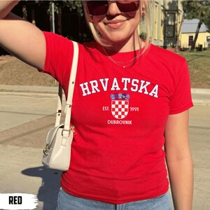 May include: A red t-shirt with the word "HRVATSKA" in white arched text. Below the text is a crest with a checkered pattern and the text "EST. 1991 DUBROVNIK". The shirt is short-sleeved.