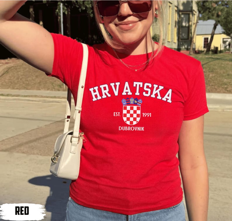 May include: A red t-shirt with the word "HRVATSKA" in white arched text. Below the text is a crest with a checkered pattern and the text "EST. 1991 DUBROVNIK". The shirt is short-sleeved.