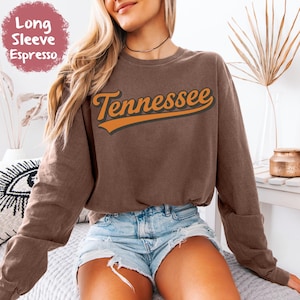 May include: Espresso-colored long-sleeve sweatshirt with the word "Tennessee" in orange script. The sweatshirt has a crew neck and long sleeves. The text "Long Sleeve Espresso" is in a circular label in the upper left corner.