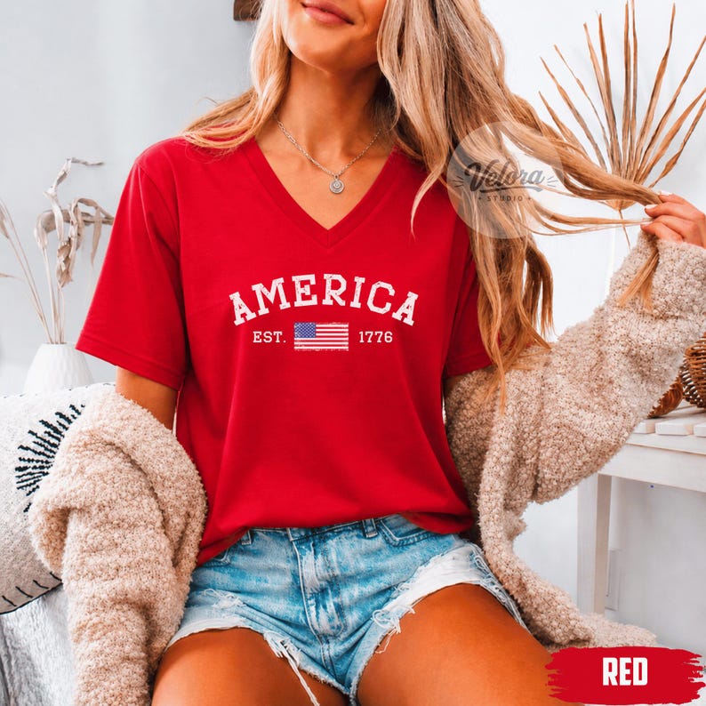May include: A red V-neck t-shirt with the word "AMERICA" in white block letters, the American flag, and the year "1776". The shirt is part of a patriotic collection.