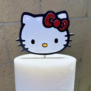 May include: A white Hello Kitty cake topper with a black outline and a red glitter bow. The topper is attached to a white foam base, perfect for decorating a cake or other dessert.