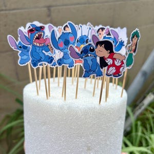 Puede incluir: Un conjunto de adornos para cupcakes del personaje Stitch azul sobre palitos de madera. Los adornos muestran a Stitch en varias poses, incluyendo uno besando a Lilo. Est&aacute;n dispuestos sobre una base de espuma blanca.