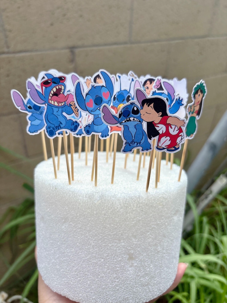 Puede incluir: Un conjunto de adornos para cupcakes del personaje Stitch azul sobre palitos de madera. Los adornos muestran a Stitch en varias poses, incluyendo uno besando a Lilo. Est&aacute;n dispuestos sobre una base de espuma blanca.