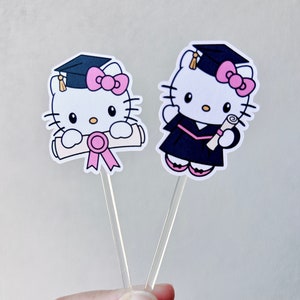 May include: Two graduation themed Hello Kitty cake toppers. The toppers are white with pink bows and black eyes. One topper has a black graduation cap and gown and is holding a diploma. The other topper has a black graduation cap and is holding a diploma with a pink ribbon.