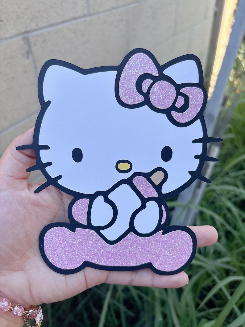 Puede incluir: Una figura de Hello Kitty blanca y rosa con un contorno negro. El lazo y la base son de color rosa brillante. La figura sostiene un biber&oacute;n y tiene ojos y bigotes negros.