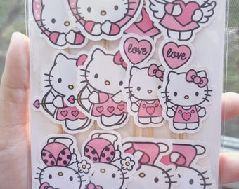May include: A set of 10 pink and white Hello Kitty cupcake toppers. The toppers feature Hello Kitty in various poses, including holding a heart, wearing angel wings, and dressed as a ladybug. The toppers are printed on cardstock and have a stick attached to the back.