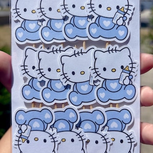 Puede incluir: Un conjunto de doce adornos de papel troquelado para cupcakes con el personaje Hello Kitty en azul claro, vistiendo un conjunto azul con un coraz&oacute;n blanco y sosteniendo un biber&oacute;n. Cada adorno est&aacute; sujeto a un palillo de madera.