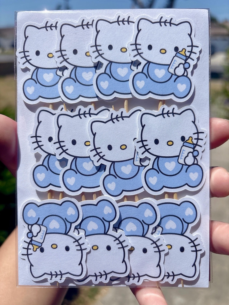 Puede incluir: Un conjunto de doce adornos de papel troquelado para cupcakes con el personaje Hello Kitty en azul claro, vistiendo un conjunto azul con un coraz&oacute;n blanco y sosteniendo un biber&oacute;n. Cada adorno est&aacute; sujeto a un palillo de madera.