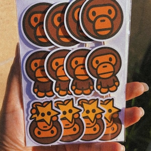 May include: A sheet of stickers featuring cartoon monkey and star designs. The stickers are in shades of brown and orange, with a white border. The stickers are held in a person's hand.