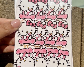 May include: A set of twelve Hello Kitty cupcake toppers arranged in three rows on a white backing card. Each topper features a white Hello Kitty character with a pink bow, sitting on a pink base, attached to a thin wooden toothpick for food use.