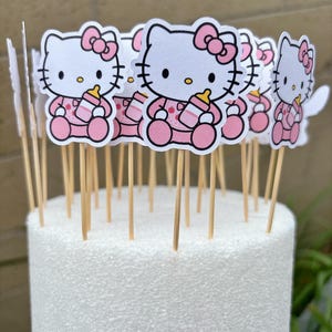Puede incluir: Una colecci&oacute;n de adornos para cupcakes de Hello Kitty que muestran al personaje con un conjunto rosa y un lazo a juego, sosteniendo un biber&oacute;n. Cada figura est&aacute; pegada a un palillo de madera fino, dise&ntilde;ado para insertarse en un pastel.