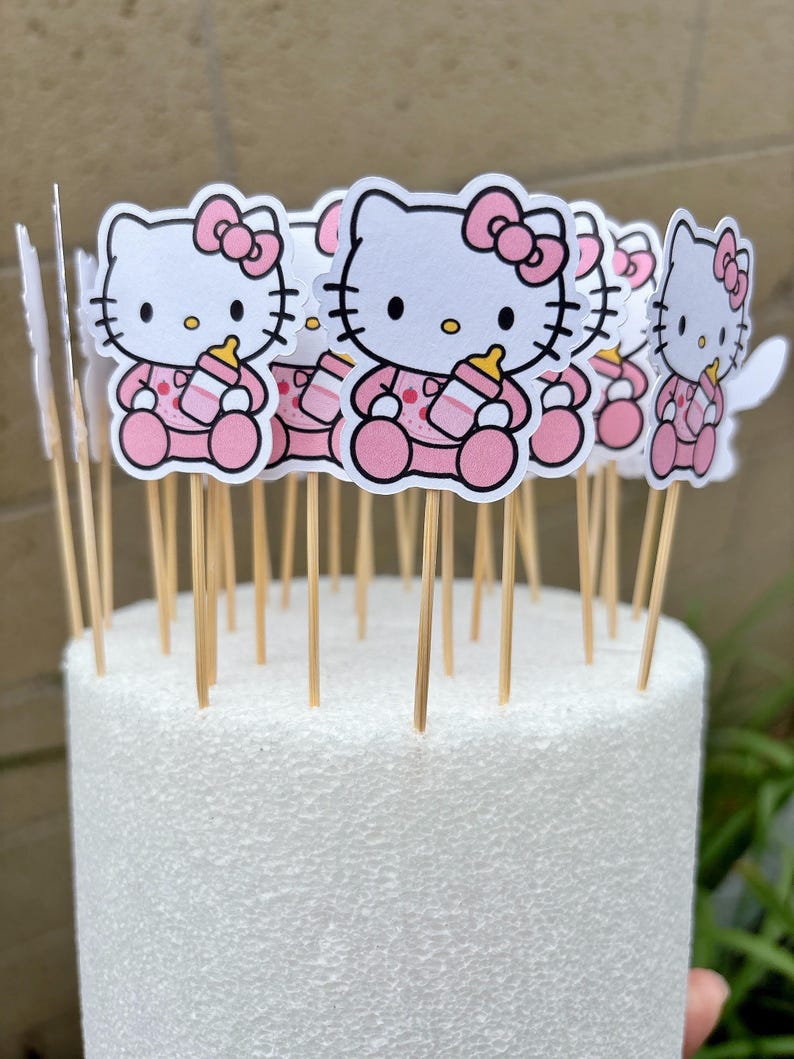 Puede incluir: Una colecci&oacute;n de adornos para cupcakes de Hello Kitty que muestran al personaje con un conjunto rosa y un lazo a juego, sosteniendo un biber&oacute;n. Cada figura est&aacute; pegada a un palillo de madera fino, dise&ntilde;ado para insertarse en un pastel.