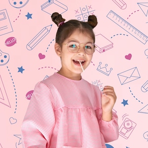 Könnte beinhalten: Ein junges Mädchen trägt eine rosa Brille und ein rosa-weiß kariertes Kleid mit einer Tasche. Sie hält einen Lolli in der Hand und lächelt. Der Hintergrund ist rosa mit verschiedenen schulischen Zeichnungen.