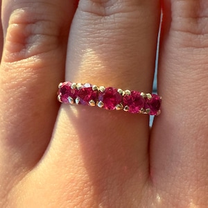 May include: A gold ring featuring a line of bright red gemstones. The round-cut stones are set closely together in a gold band. The ring is displayed on a finger, highlighting the gemstones' colour and sparkle. The design is classic.