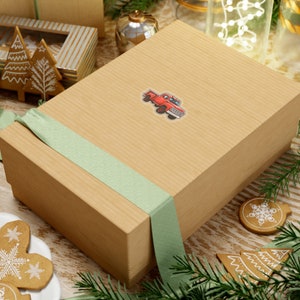 May include: A brown gift box with a green ribbon and a red truck sticker. The box is sitting on a wooden surface with a few gingerbread cookies and a pine branch.