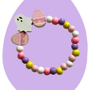 May include: A colorful wooden Easter bracelet with pink, yellow, white, and purple beads. The bracelet features two wooden egg charms with a pink and white geometric pattern and a white wooden ghost charm.