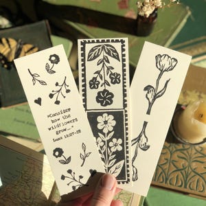 May include: Three cream-colored bookmarks with black floral designs and text. One bookmark has the quote "Consider how the wildflowers grow..." from Luke 12:27-28. The other two feature different floral illustrations. The background includes books and a small vase.