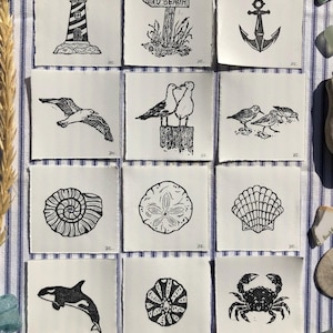 May include: A collection of twelve square white paper prints featuring black ink illustrations of coastal themes including a lighthouse, a beach sign, an anchor, seagulls, shorebirds, a spiral shell, a sand dollar, a scallop shell, an orca, a sea urchin, and a crab.
