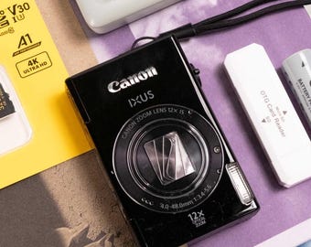 Może przedstawiać: Czarny aparat cyfrowy Canon IXUS, szara ładowarka do akumulatorów Canon z czarnym kablem, karta pamięci microSDHC Kodak 32GB w żółtym opakowaniu, biały czytnik kart OTG oraz szary akumulator Canon NB-8L ułożone na płaskiej powierzchni.