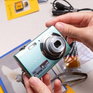 Op de afbeelding: Een lichtblauwe Nikon Coolpix digitale camera met een 3x optische zoomlens, een 2,5-inch LCD-scherm en 10 megapixels. De voorkant van de camera toont de merknaam Nikon en het model Coolpix met de bijbehorende technische specificaties.