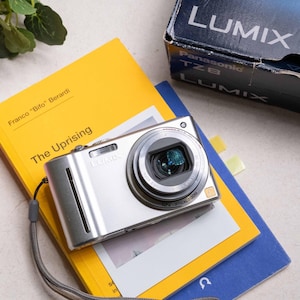 Op de afbeelding: Een zilverkleurige Panasonic Lumix TZ8 digitale camera met een 12x optische zoomlens en een grijze polsband, rustend op een geel boek getiteld The Uprising van Franco Bifo Berardi, naast een donkerblauwe Panasonic Lumix TZ8 cameradoos.