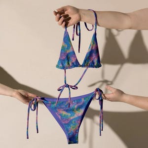 Op de afbeelding: Een tweedelige bikiniset met een levendig, wervelend marmerpatroon in blauwe, paarse, roze en gele tinten. De top heeft driehoekige cups met dunne strikbandjes en het broekje is voorzien van verstelbare zijstrikjes met een paarse rand.
