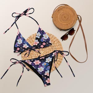 May include: A two-piece bikini set featuring a dark blue background with a pattern of pink and white cherry blossoms and clouds. The top has thin halter straps and the bottom has side ties. A round woven straw bag and brown sunglasses are included.