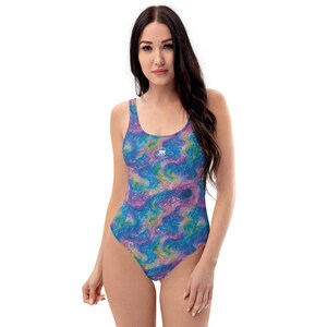 Peut inclure: Un maillot de bain une pièce arborant un motif abstrait tourbillonnant aux tons bleu, violet et jaune. Le vêtement présente une encolure dégagée, de larges bretelles et une coupe échancrée, avec un petit logo blanc centré sur la poitrine.