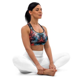 Puede incluir: Sujetador deportivo colorido con un diseño de nubes en tonos rojos, azules y negros. Se combina con leggings blancos. La persona está sentada con las piernas cruzadas, con los ojos cerrados, sugiriendo yoga o fitness.