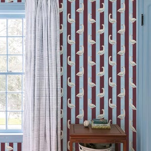May include: Wallpaper featuring a repeating pattern of white geese on alternating vertical stripes of light blue and dark maroon. A light gray textured curtain hangs beside a wooden side table with a striped fabric basket underneath.