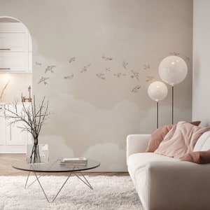 May include: A white couch with two pink throw pillows sits in a living room with a white wall decorated with a flock of small brown birds. Two white globe lamps stand on either side of the couch. A glass coffee table with a gold metal frame sits in front of the couch.