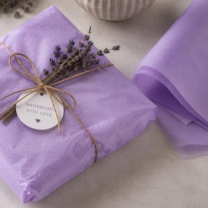May include: A square gift package wrapped in light purple tissue paper, tied with brown twine. A sprig of dried lavender is tucked under the twine, alongside a round white tag featuring the text HANDMADE WITH LOVE and a small heart icon.