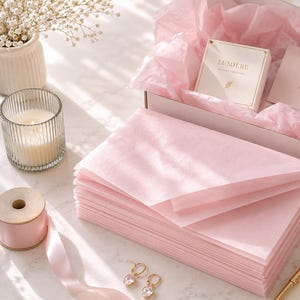 Op de afbeelding: Een stapel lichtroze vloeipapier in een witte geschenkdoos bekleed met bijpassend roze vloeipapier. De doos bevat een klein wit doosje met het label LUMIÈRE en een bedankkaartje. Accessoires zijn onder meer een kaars, lint en sieraden.