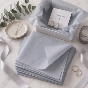 May include: A stack of light gray tissue paper sheets next to a white gift box containing two silver hoop earrings and a card with the text THANK YOU. A silver necklace rests on a small scalloped dish, and two silver rings lie nearby with ribbon.