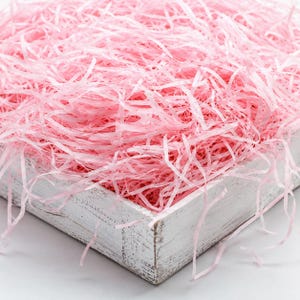 Pode incluir: Uma caixa de madeira branca com acabamento desgastado preenchida com uma grande quantidade de papel picado rosa claro. As tiras finas de papel amassado estão empilhadas, criando uma camada texturizada e volumosa que transborda pelas bordas.