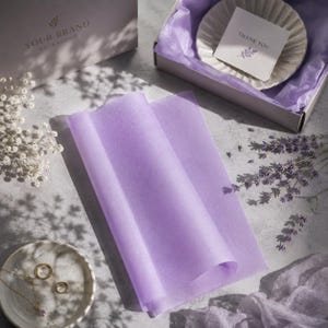 Può includere: Un foglio di carta velina viola chiaro piegato su una superficie, accompagnato da una scatola regalo contenente un piattino bianco smerlato con un biglietto di ringraziamento, due anelli d'oro su un piattino, lavanda secca e fiori bianchi.