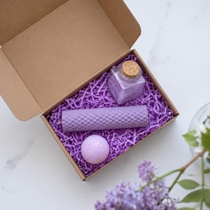 Pode incluir: Uma caixa de presente de papelão marrom preenchida com papel picado roxo, contendo um pote de vidro quadrado com tampa de cortiça cheio de sais de banho roxos, uma vela cilíndrica de cera de abelha com textura de favo de mel e uma bomba de banho roxa.