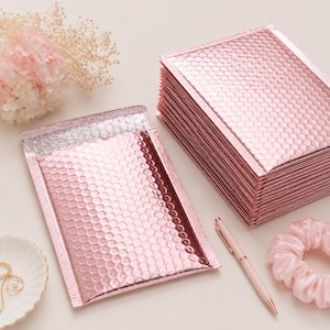 May include: A stack of pink bubble mailers with a metallic finish. The mailers are next to a single mailer, a pink pen, and a pink scrunchie. The mailers have a bubble wrap texture. The background is a light pink.