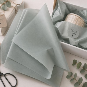 Peut inclure: Une pile de feuilles de papier de soie vert sauge à côté d'une boîte cadeau blanche contenant une bougie beige enveloppée dans du papier de soie assorti avec un autocollant rond portant l'inscription thank you et un cœur, de la ficelle et des ciseaux noirs.