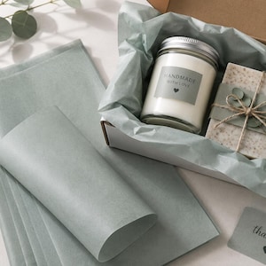 Peut inclure: Un coffret cadeau contenant une bougie blanche dans un pot en verre avec une étiquette HANDMADE WITH LOVE et un cœur, et un savon rectangulaire moucheté enveloppé de papier vert sauge et de ficelle, avec du papier de soie et une étiquette.