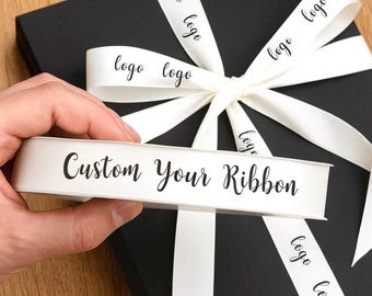 May include: A black gift box tied with a white ribbon featuring the word "logo" printed repeatedly. A white ribbon roll with the text "Custom Your Ribbon" is held in front of the box.