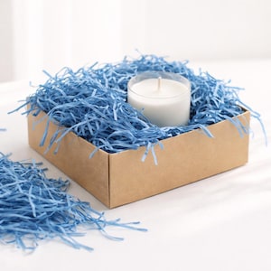 Può includere: Una candela bianca in un supporto di plastica trasparente è posta in una scatola di cartone marrone riempita di carta blu sminuzzata. La scatola è su una superficie bianca, con più carta blu sparsa.