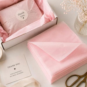 May include: A white cardboard box filled with light pink tissue paper and a thank you sticker, a stack of folded light pink tissue paper, a small thank you card, gold-toned scissors, and a small ceramic dish with gold rings.