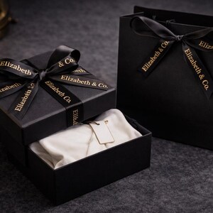 May include: Black gift box with a satin ribbon and the text "Elizabeth & Co." in gold. The box is open, revealing a white fabric and a tag. A matching black gift bag with a ribbon is also present.